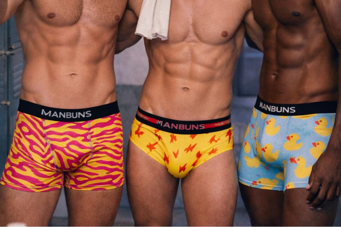 Three men wearing colorful underwear with 'Manbuns' branding in a locker room setting.
